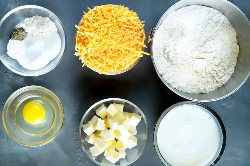 Overhead horizontal image of glass and stainless steel bowls of dry seasonings, egg, shredded cheese, cubed butter, flour, and buttermilk, on a gray surface.