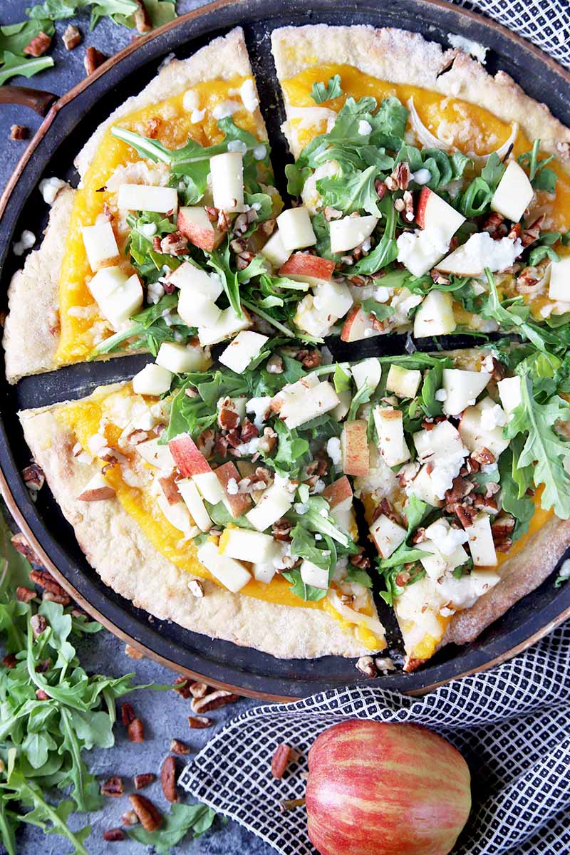 Vertical overhead image of a homemade pizza on a metal baking pan that has been sliced into four pieces, on a gray surface with a black and white checkered cloth, a whole apple, and scattered fresh arugula and chopped nuts.