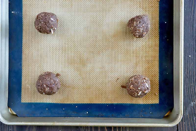 Horizontal overhead image of four balls of uncooked chocolate dough on a blue and beige silicone pan liner set into a metal rimmed baking sheet, on a gray surface.