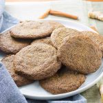 Horizontal image of a white ceramic plate of homemade gluten-free buckwheat ginger cookies, on a gray surface with scattered whole cinnamon sticks, a folded pale blue cloth, fresh ginger, and another smaller plate of the same dessert to the right of the frame.