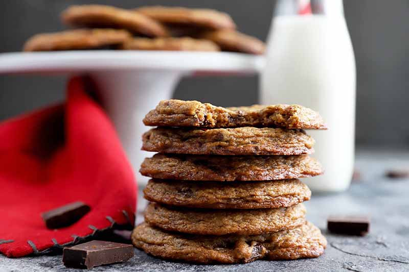 Horizontal image of a stack of thin, dark cookies next to a red towel and white cake stand.