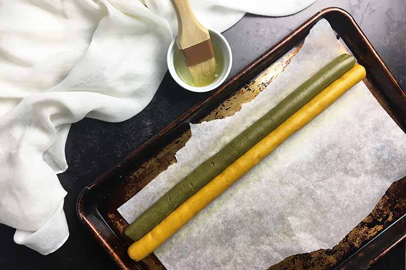 Horizontal image of a yellow and a green long log of dough on a baking sheet next to a dish with a brush and egg whites.