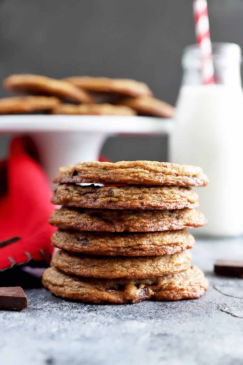 Vertical image of a stack of thin, dark brown chocolate chip cookies in front of a white cake stand, red towel, and glass of milk. 