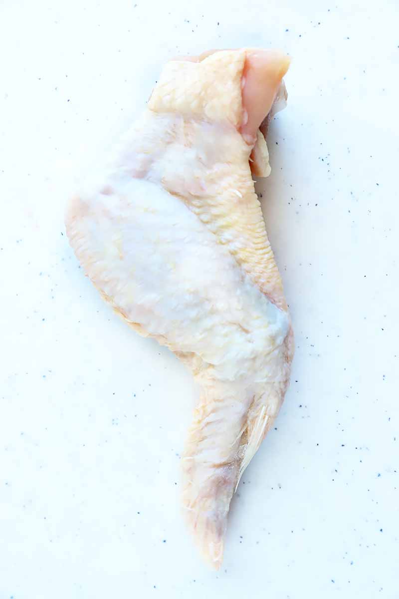 Vertical image of one whole raw chicken wing on a white cutting board.