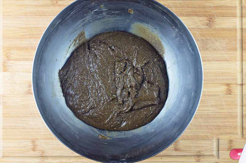 Overhead horizontal image of a dark brown batter in a stainless steel mixing bowl, on a beige wood surface.