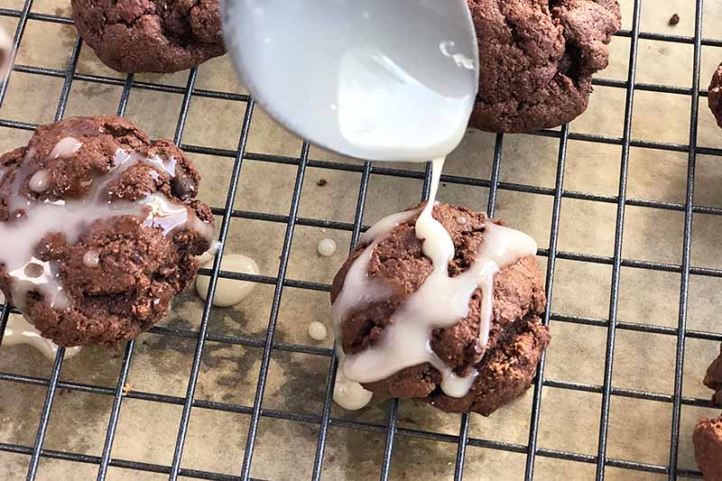 Horizontal closely cropped overhead image of a spoon dripping a white glaze onto round chocolate cookies on a cooling rack below, with light brown parchment paper underneath.