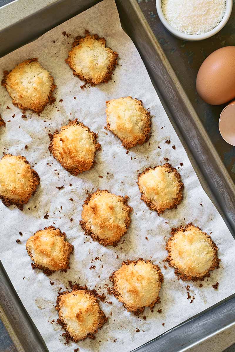 Vertical closely cropped overhead image of golden brown baked macaroons on a metal baking sheet topped with parchment paper, on a dark gray countertop with brown eggs and a ceramic cup of flour.