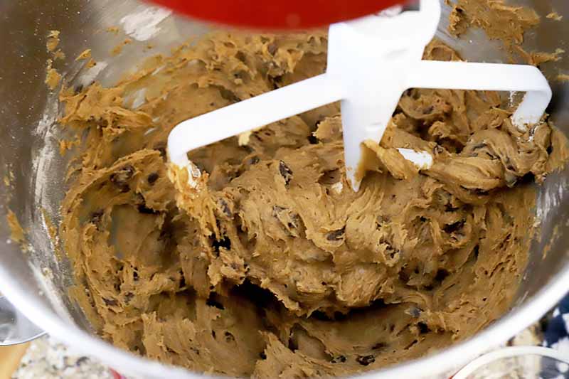 Horizontal image of a thick dough with chocolate chips in the bowl of a stand mixture fitted with the paddle attachment.