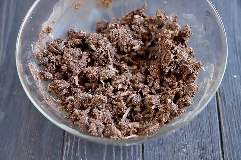 Horizontal closely cropped overhead image of a brown chocolate dough flecked with coconut, in a large mixing bowl on a dark brown surface.