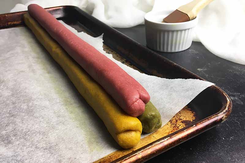 Horizontal image of three logs of colored dough stacked to form a triangle on a baking sheet next to a dish with a pastry brush.