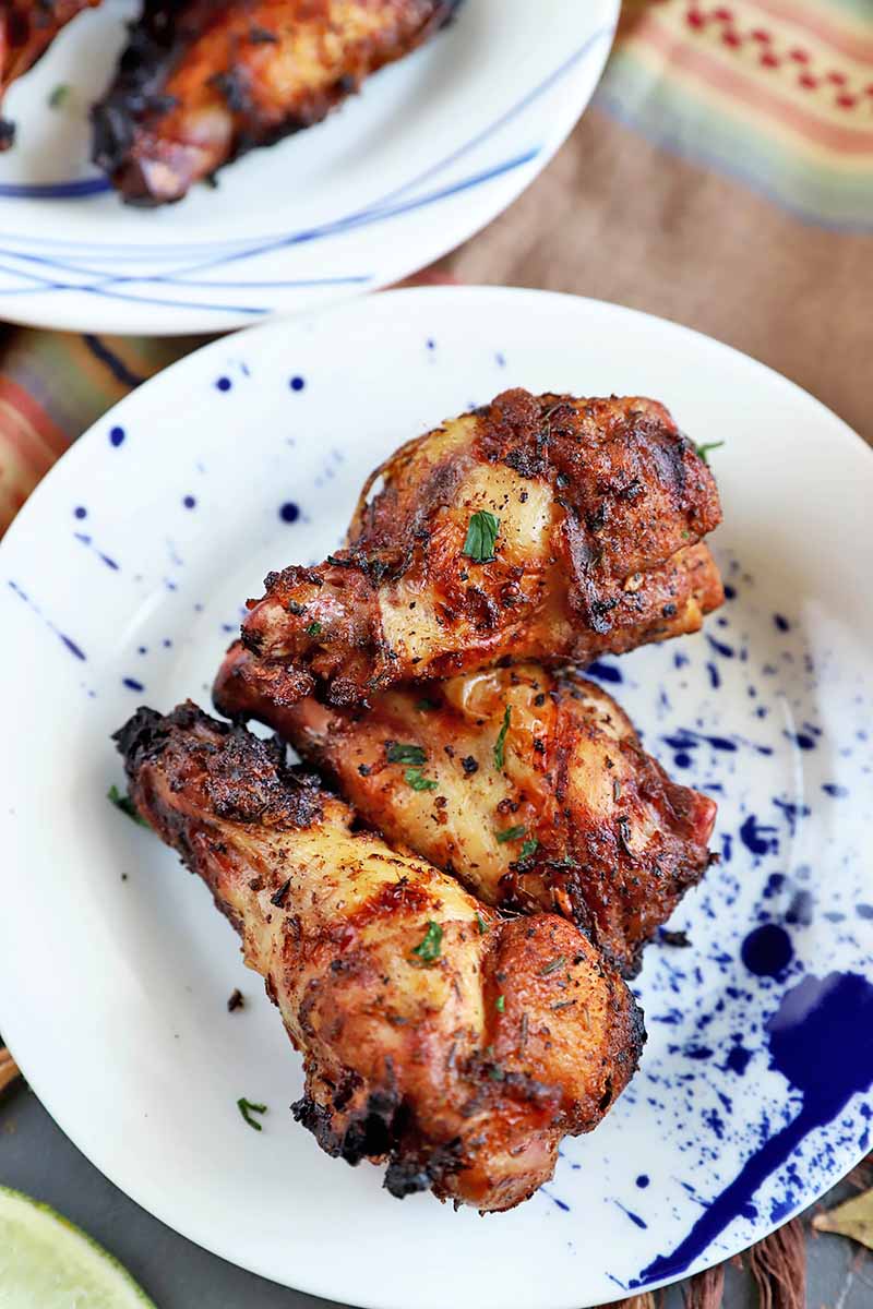 Vertical image of three grilled chicken wings with a Jamaican jerk marinade, on a white plate with a blue splatter pattern, beside another plate on a brown surface.