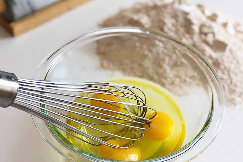Horizontal image of a whisk mixing raw eggs in a glass bowl in front of a mound of flour.
