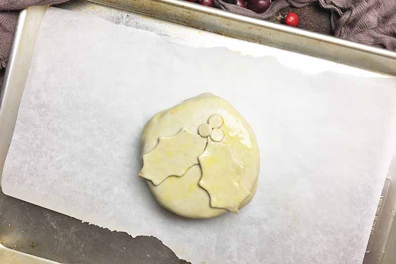 Horizontal image of a baking sheet with parchment paper and an unbaked pastry decorated with holly-shaped dough.