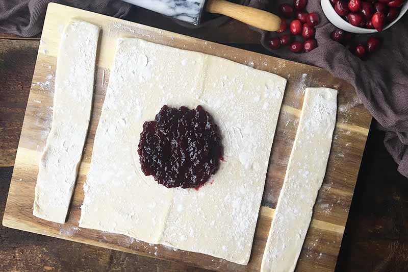 Horizontal image of jam spread in the center of a square piece of dough with scraps on the sides on a wooden board.