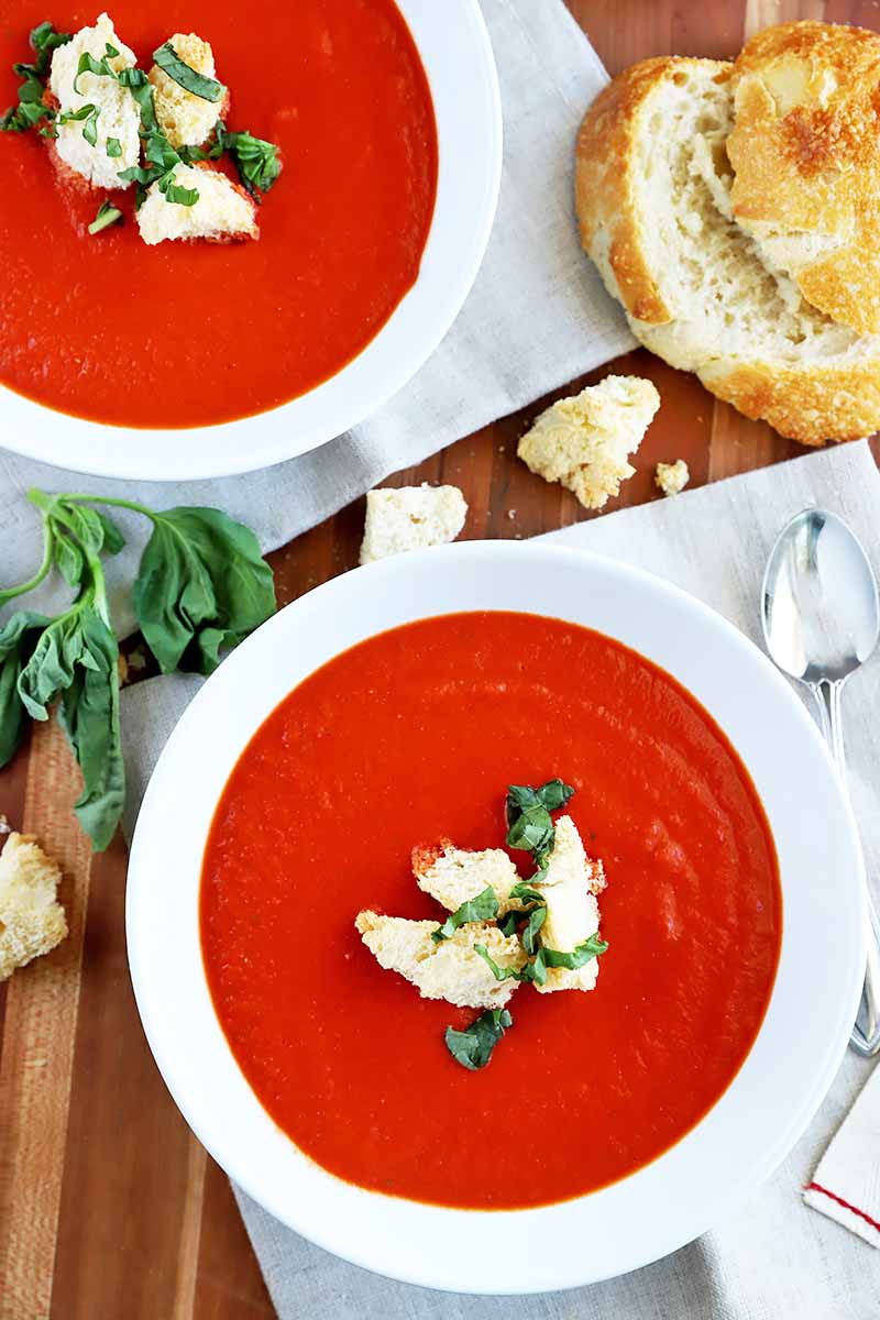 Vertical overhead image of two white ceramic bowls filled with tomato soup, garnished with toasted bread cubes and chopped basil, on a brown wood surface with slices and pieces of bread and a sprig of fresh herbs, on pale gray cloth placemats.