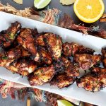 Horizontal overhead image of a white rectangular ceramic platter of homemade grilled wings, on a gray surface on a multicolored cloth with fringe, with scattered pieces of cut citrus, bay leaves, and whole cinnamon sticks.