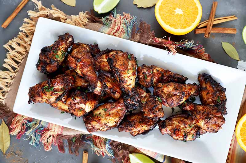 Horizontal overhead image of a white rectangular ceramic platter of homemade grilled wings, on a gray surface on a multicolored cloth with fringe, with scattered pieces of cut citrus, bay leaves, and whole cinnamon sticks.