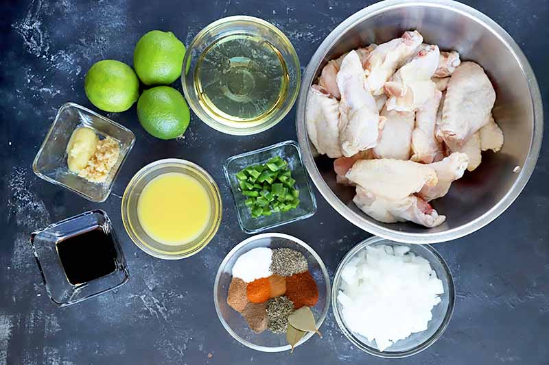 Overhead horizontal image of square and round glass and stainless steel bowls of various sizes, filled with soy sauce, minced ginger and garlic, orange juice, oil, diced jalapeno, chopped onion, raw chicken wings, and various spices, with three whole limes, on a mottled gray surface.