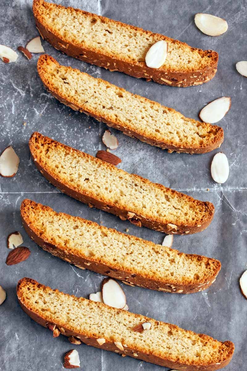 Vertical overhead image of five just-baked biscotti on a sheet of waxed paper with scattered sliced almonds.