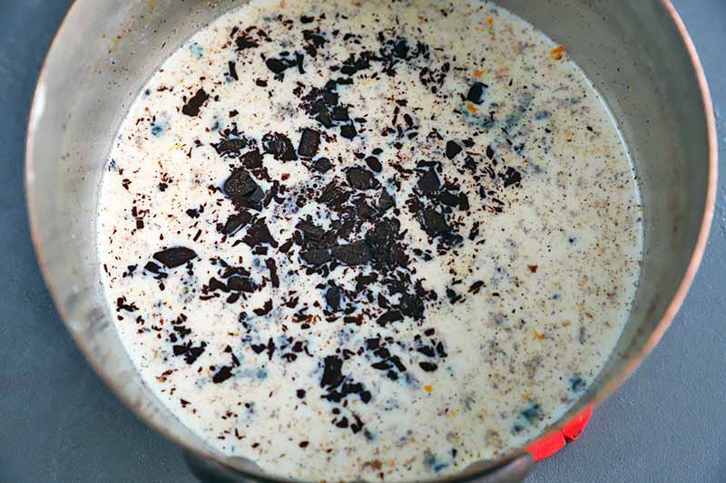 Overhead closely cropped horizontal image of a saucepan filled with a milk mixture flecked with orange zest and pieces of finely chopped chocolate, on a gray surface.