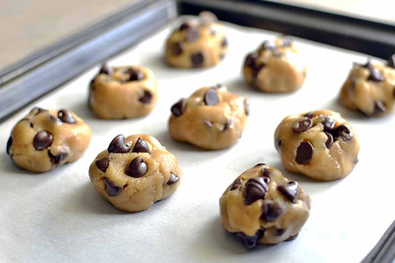 Horizontal image of nine roughly spherical portions of chocolate chip cookie dough of equal size arranged in three rows on a parchment-lined rimmed metal baking sheet, on a beige background.