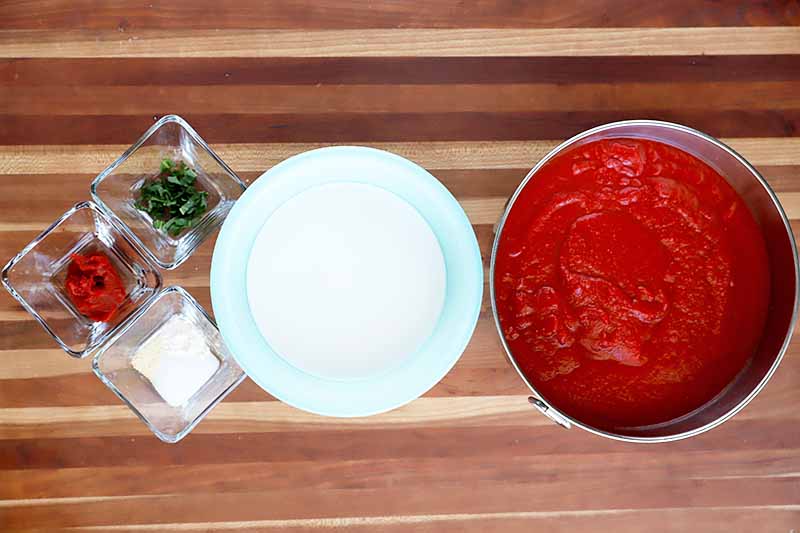 Horizontal overhead image of three small square glass bowls of chopped basil, tomato paste, and spices, a larger pale blue bowl of coconut milk, and a stainless steel bowl of pureed canned tomatoes, on a striped brown and blonde wood surface.