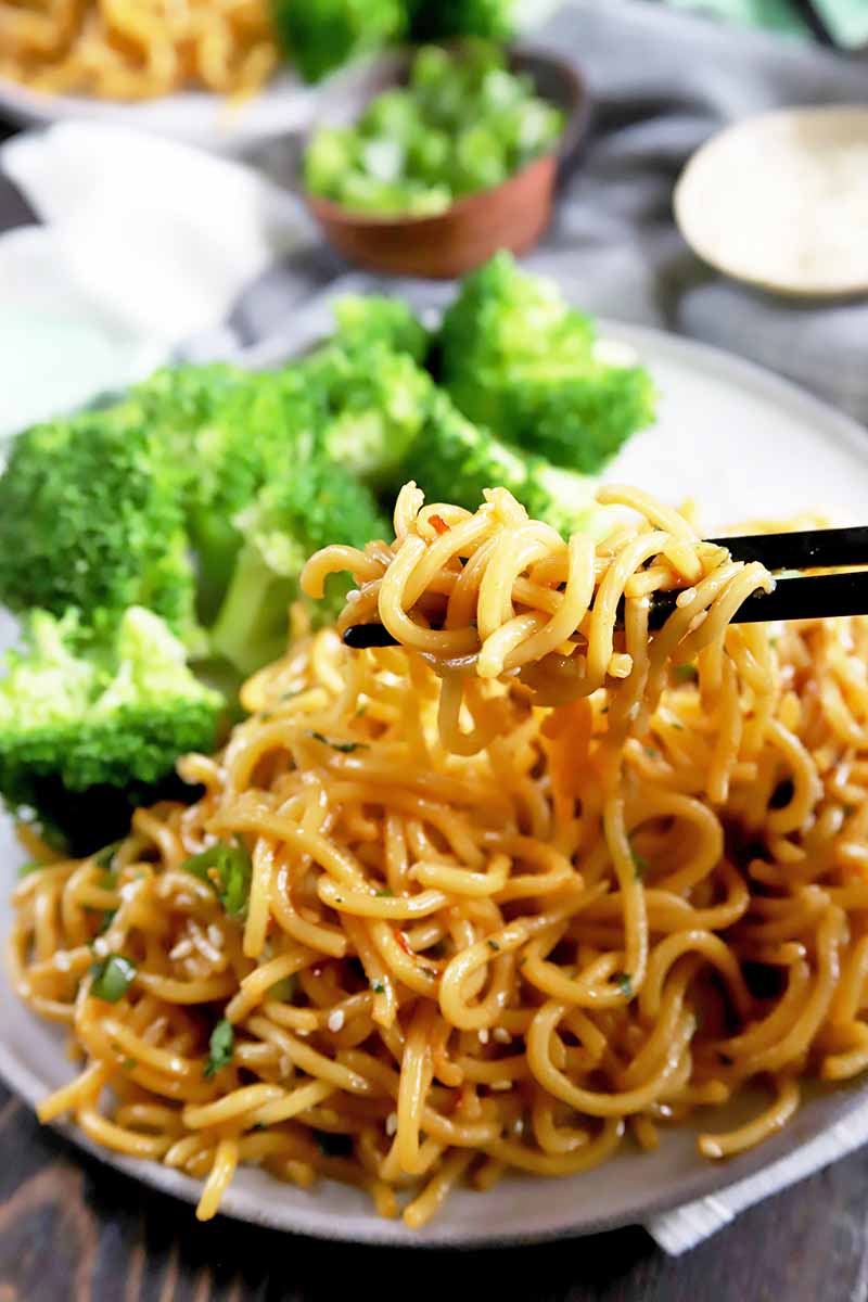 Vertical image of chopsticks picking up strands of pasta on a plate with broccoli.