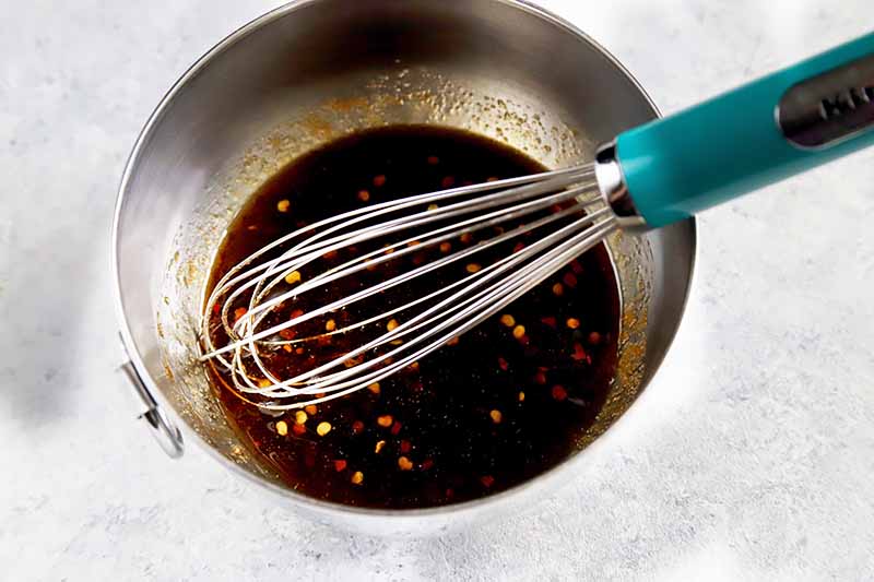 Horizontal image of a blue whisk in a metal bowl with soy sauce and other seasonings.
