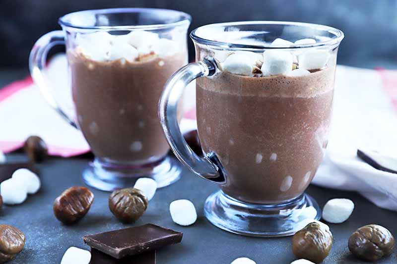 Horizontal image of two clear glass mugs with handles, filled with homemade hot chocolate, on a gray surface with scattered pieces of a dark chocolate bar, whole roasted chestnuts, and mini marshmallows, with a white kitchen towel with red trim in the background in soft focus.
