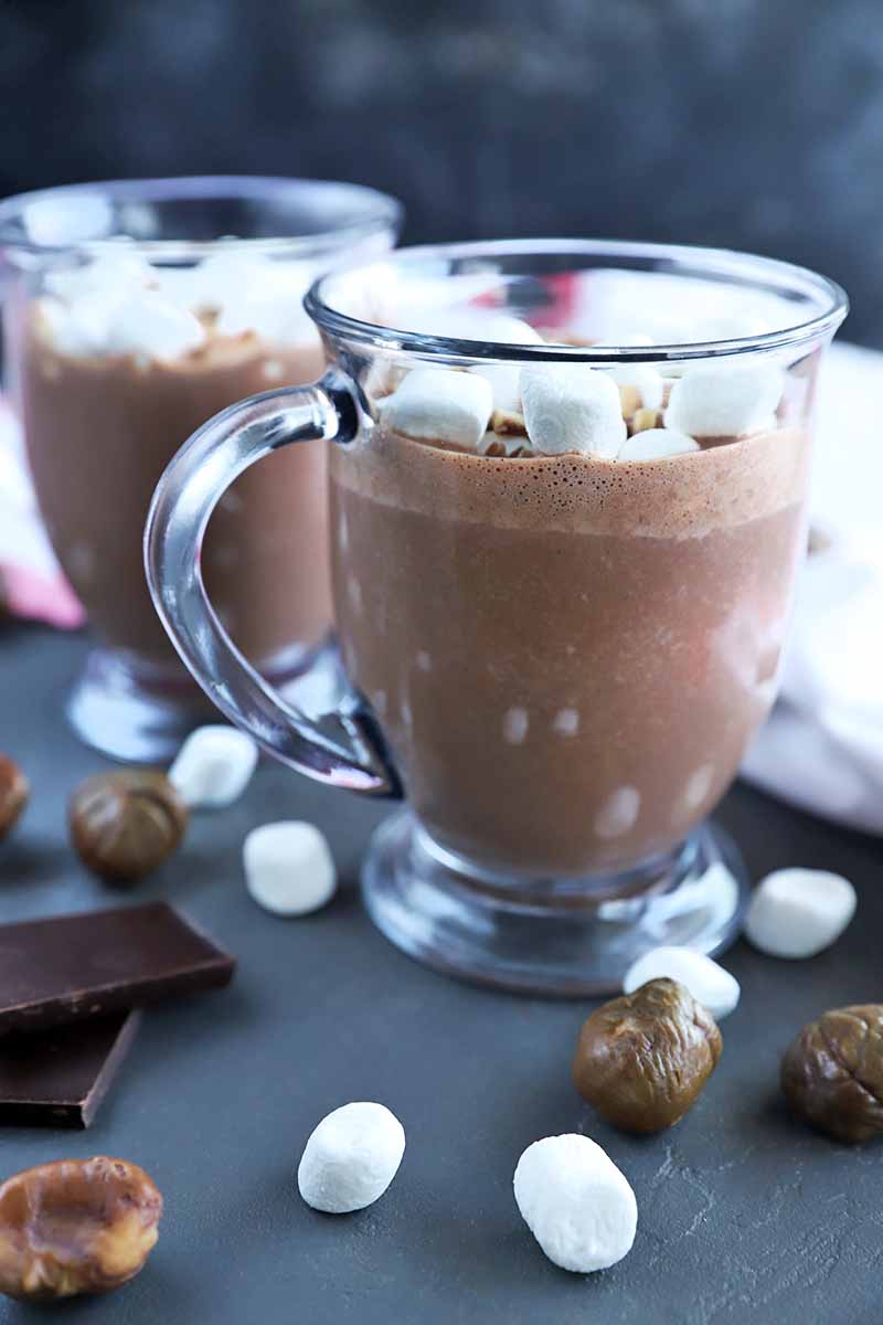 Vertical image of two clear glass mugs of hot chocolate, on a gray surface with scattered mini marshmallows, whole roasted chestnuts, and pieces of dark chocolate, with a white kitchen towel with red trim to the right, against a mottled dark gray background.