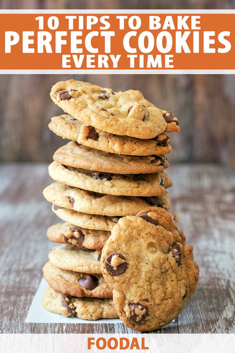 Vertical image of a stack of ten homemade chocolate chip cookies with another propped up in front of the stack, on a small rectangle of parchment paper on top of a weathered whitewashed wood surface, printed with orange and white text at the top and bottom of the frame.