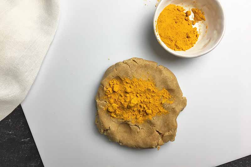 Horizontal image of a disc of tan dough topped with turmeric powder next to a dish with more turmeric.