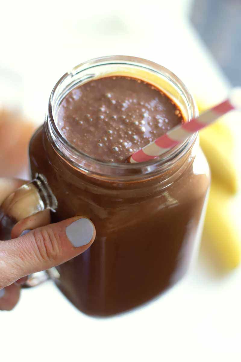 Vertical image of a hand holding a glass mug with a creamy chocolate beverage and a straw.