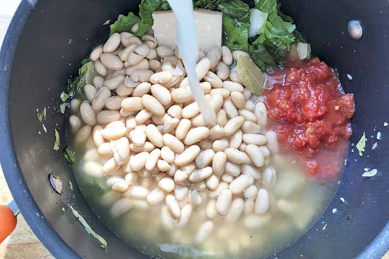 Horizontal image of cannellini, tomatoes, escarole, and broth in a large pot.