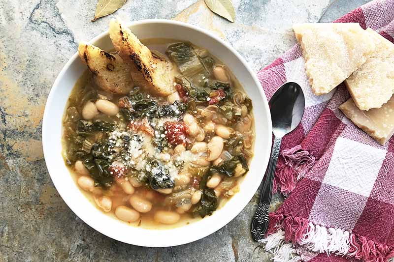 Horizontal image of a bowl of soup with cannellini and escarole on a red checkered towel.