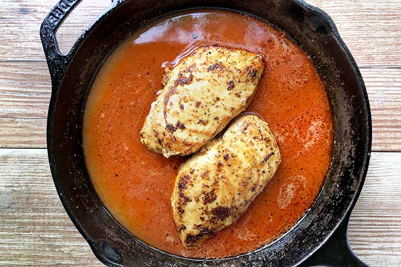 Horizontal overhead image of a cast iron pan of Buffalo sauce and seasoned chicken breasts, on a wood background.