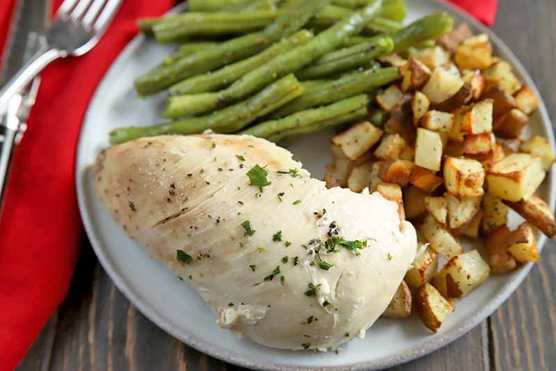 Horizontal overhead image of a slow cooked skinless chicken breast coated with herbs, on a plate with green beans and roasted potatoes, on a brown wood surface with a folded red cloth napkin topped with a fork to the left of the frame.