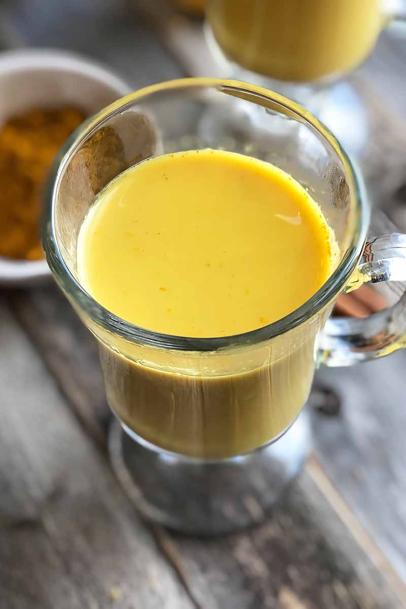 Vertical image of a glass mug filled with a turmeric-infused beverage on a wooden table in front of a bowl of ground cinnamon.