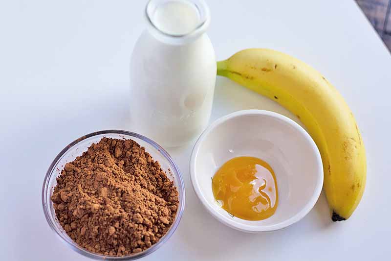 Horizontal image of a bowl of cacao powder, a bowl of honey, a glass of kefir, and a banana.