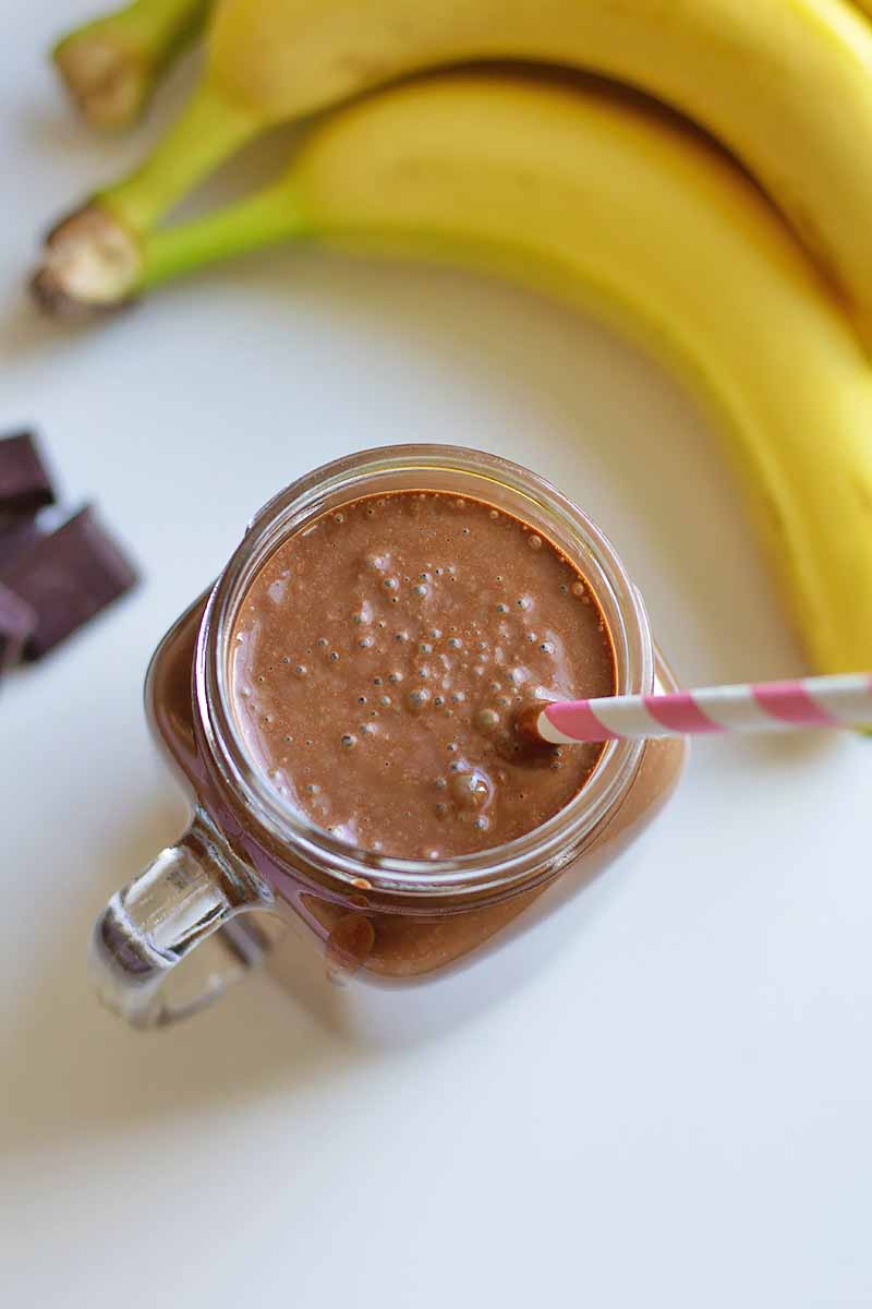 Vertical top-down image of a dark brown smoothie in a glass mug with a straw next to bananas.