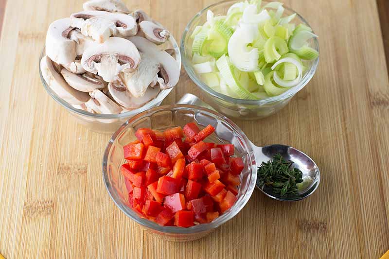 Horizontal image of sliced mushrooms, leeks, peppers, and herbs in bowls on a wooden board.