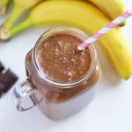 Horizontal image of a glass mug with a cacao smoothie and straw next to whole bananas and chocolate pieces.