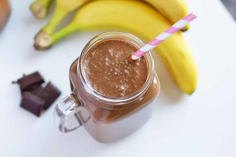 Horizontal image of a glass mug with a cacao smoothie and straw next to whole bananas and chocolate pieces.