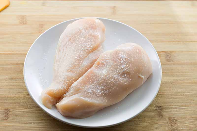 Horizontal image of two raw chicken breasts with salt on a white plate.