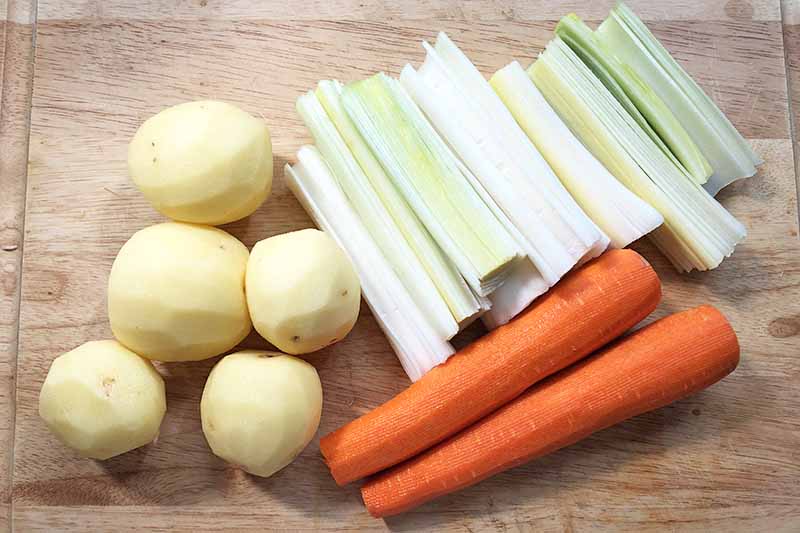 Horizontal image of prepped potatoes, carrots, and leeks on a wooden surface.