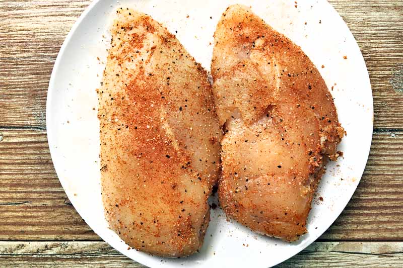 Horizontal overhead image of two raw seasoned skinless and boneless chicken breasts on a white plate, on a wood background.