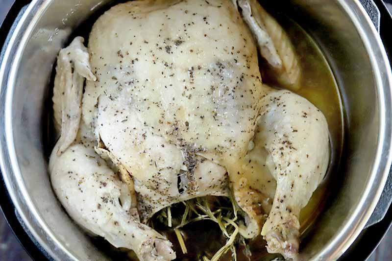 Horizontal overhead image of a whole cooked chicken filled with fresh herbs and sprinkled with black pepper, in a metal slow cooker insert with about a cup of broth.