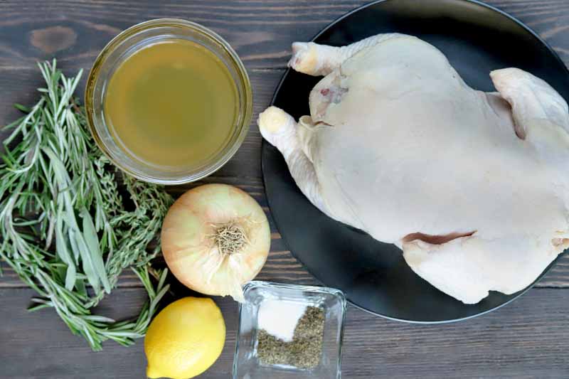 Horizontal overhead image of springs of fresh herbs, a yellow onion, a lemon, a small square glass dish of salt and pepper, a small round glass dish of chicken broth, and a raw whole chicken on a black ceramic plate, on a brown wood table.