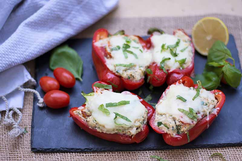 Horizontal image of four stuffed red peppers on a slate next to tomatoes and fresh herbs.