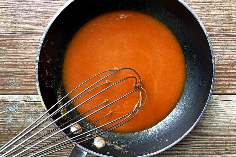 Overhead horizontal image of a whisk stirring red hot sauce in a nonstick frying pan, on a brown wood surface.
